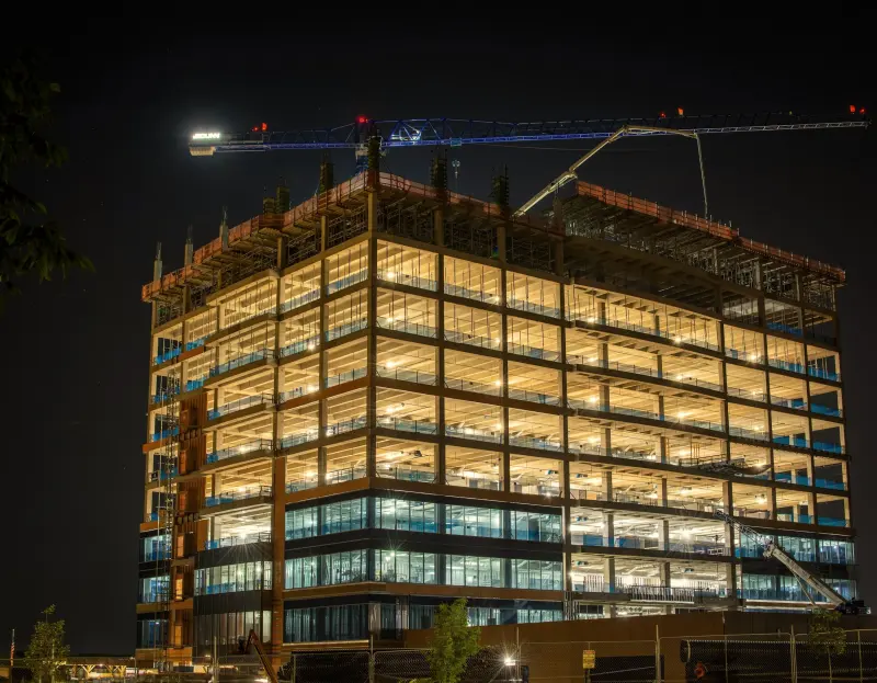 A multi-storey commercial building under construction, its floors lit at night beneath a tower crane.